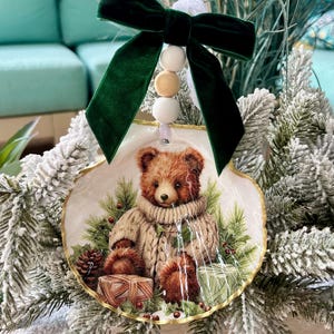 May include: A decorative Christmas ornament featuring a teddy bear wearing a sweater, set against a backdrop of pine needles and wrapped gifts. The ornament is adorned with a green velvet bow and wooden beads, creating a festive holiday decoration.