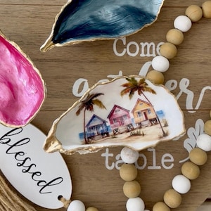 May include: Oyster shell trinket dishes in pink, blue, and a shell with a beach scene. The shells have gold painted edges. A wooden bead garland and a white tag with the word "blessed" are also in the image.