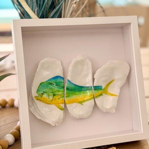 May include: A white framed artwork featuring three oyster shells, each painted with a colorful fish design. The fish are depicted with blue, green, and yellow hues, creating a coastal theme. The artwork is displayed on a wooden surface.