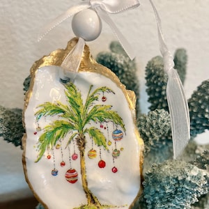 May include: Hand-painted oyster shell Christmas ornament. The white shell features a palm tree with colorful ornaments, a gold-painted edge, and a white ribbon for hanging. A white bead is attached to the ribbon.