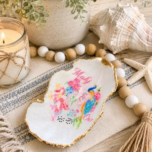 May include: A white oyster shell trinket dish with a gold-painted edge, featuring a colorful graphic of a dragon, a peacock, flowers, and dice, with the text "Mahj On!" printed in pink script across the center of the smooth, concave surface.