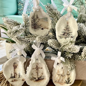 May include: Set of five oyster shell Christmas ornaments. Each ornament features a winter scene with a tree, lighthouse, anchor, or sailboat. The ornaments are white with glitter and have white ribbon hangers.