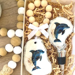 May include: A gift set with a bottle stopper and a decorative ornament, both featuring a blue dolphin design. The items are in a white box with decorative paper and wooden beads.