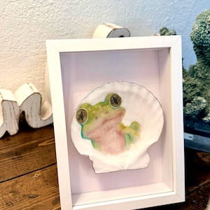 May include: A framed artwork featuring a frog painted on a seashell. The frog is green and pink with large, detailed eyes. The seashell is white, and the frame is a simple white square.