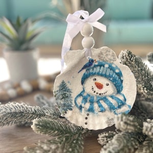 May include: A seashell ornament featuring a snowman design with a blue hat and scarf, a carrot nose, and a smiling face. The ornament is decorated with a small blue bird and a white ribbon for hanging. A festive winter decoration.