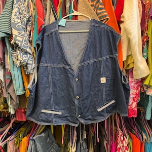 May include: A sleeveless denim vest in a medium blue wash, featuring a V-neck and button-down closure. The vest has two front pockets with white and blue striped accents and a small square patch. It hangs on a white hanger against a backdrop of colorful clothing.