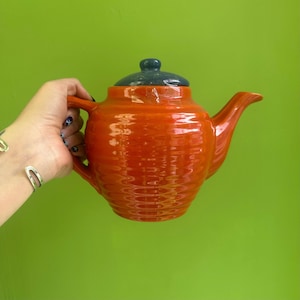 May include: An orange ceramic teapot with a textured surface and a dark blue lid. The teapot has a curved spout and handle. The background is a solid green color.