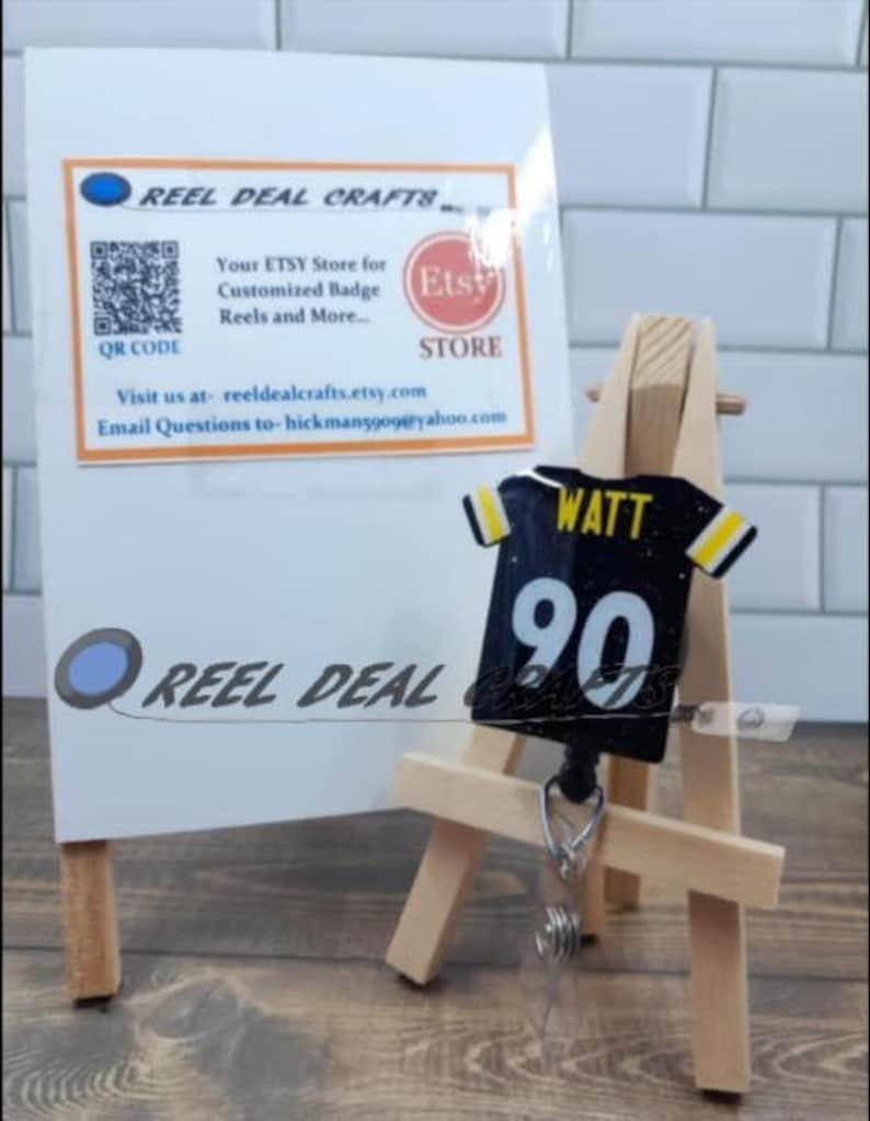 May include: A black and yellow football jersey with the name "WATT" and the number "90" on the back. The jersey is attached to a retractable badge reel and is displayed on a small wooden easel.