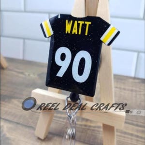 May include: Black and yellow football jersey with the name "WATT" and the number "90" on it. The jersey is attached to a retractable badge holder.