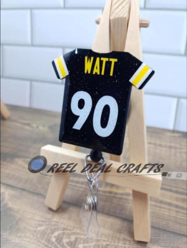 May include: Black and yellow football jersey with the name "WATT" and the number "90" on it. The jersey is attached to a retractable badge holder.