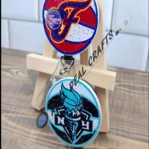 May include: Two retractable badge reels with glitter and team logos. The top reel features a red, white, and blue basketball with the word "Flyers" and a letter "F". The bottom reel features a black and white background with a blue and white torch and the letters "NY".