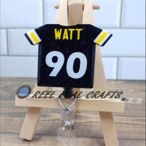 May include: Black and yellow football jersey with the name "WATT" and the number "90" on the back. The jersey is attached to a retractable badge holder.