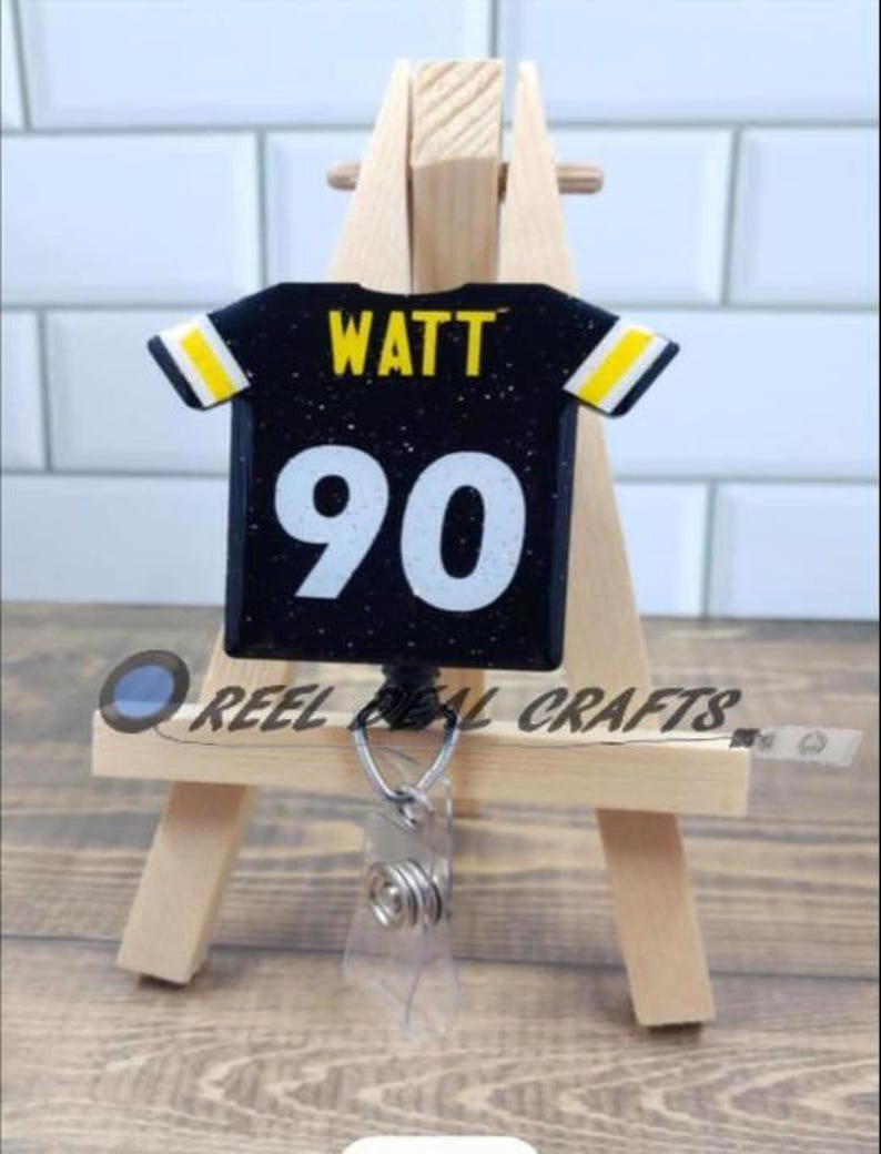 May include: Black and yellow football jersey with the name "WATT" and the number "90" on the back. The jersey is attached to a retractable badge holder.