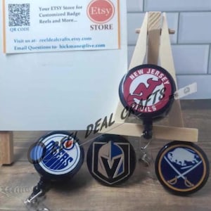 May include: A collection of customized badge reels featuring various sports team logos, including the New Jersey Devils, Edmonton Oilers, Vegas Golden Knights, and Buffalo Sabres. The reels are black with team emblems and a retractable cord.