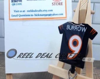 May include: A black and orange football jersey with the name "BURROW" and the number "9" on the back. The jersey is attached to a retractable badge reel with a black and white design. The reel is displayed on a small wooden easel.
