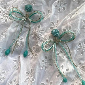 Puede incluir: Dos lazos de cuentas turquesa y oro con largas colas de cuentas. Los lazos están sobre un fondo blanco con un patrón floral.