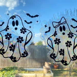 May include: Two holographic ghost window clings with black floral designs and bat accents. One ghost has a bow. The clings are displayed against a backdrop of a blue sky and trees.