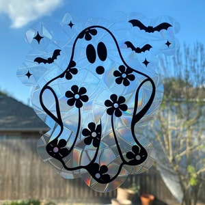 May include: A holographic ghost window cling with a floral design and bats. The ghost has black eyes and a mouth, with black flower accents. The background is a blue sky.