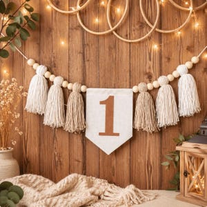 May include: A decorative garland featuring alternating white and beige yarn tassels strung on a cord with natural wooden beads. A central white fabric pennant displays the number 1 in a brown font, creating a cohesive piece for celebrations.