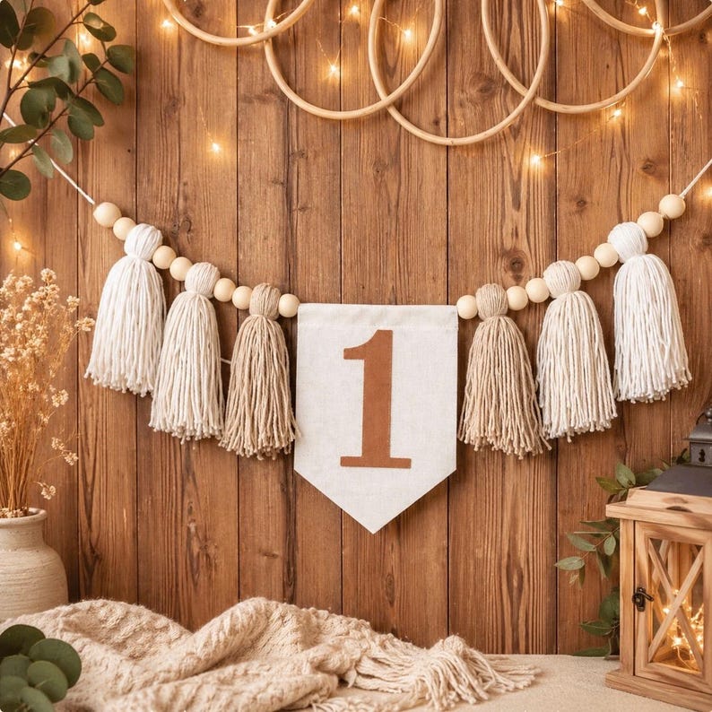 May include: A decorative garland featuring alternating white and beige yarn tassels strung on a cord with natural wooden beads. A central white fabric pennant displays the number 1 in a brown font, creating a cohesive piece for celebrations.