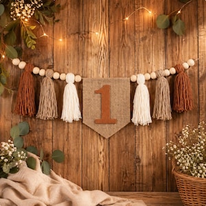 May include: A decorative banner featuring a central burlap pennant with a felt number one, flanked by wooden beads and yarn tassels in shades of rust, beige, and cream. The banner is strung horizontally with a natural, rustic aesthetic.