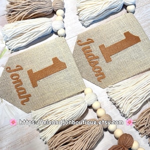 May include: Two burlap pennant banners featuring the names Jonah and Judson with the number one in brown felt. The banners are accented with strings of wooden beads and yarn tassels in shades of cream, beige, and brown. Text reads https://miaandjonboutique.etsy.com.