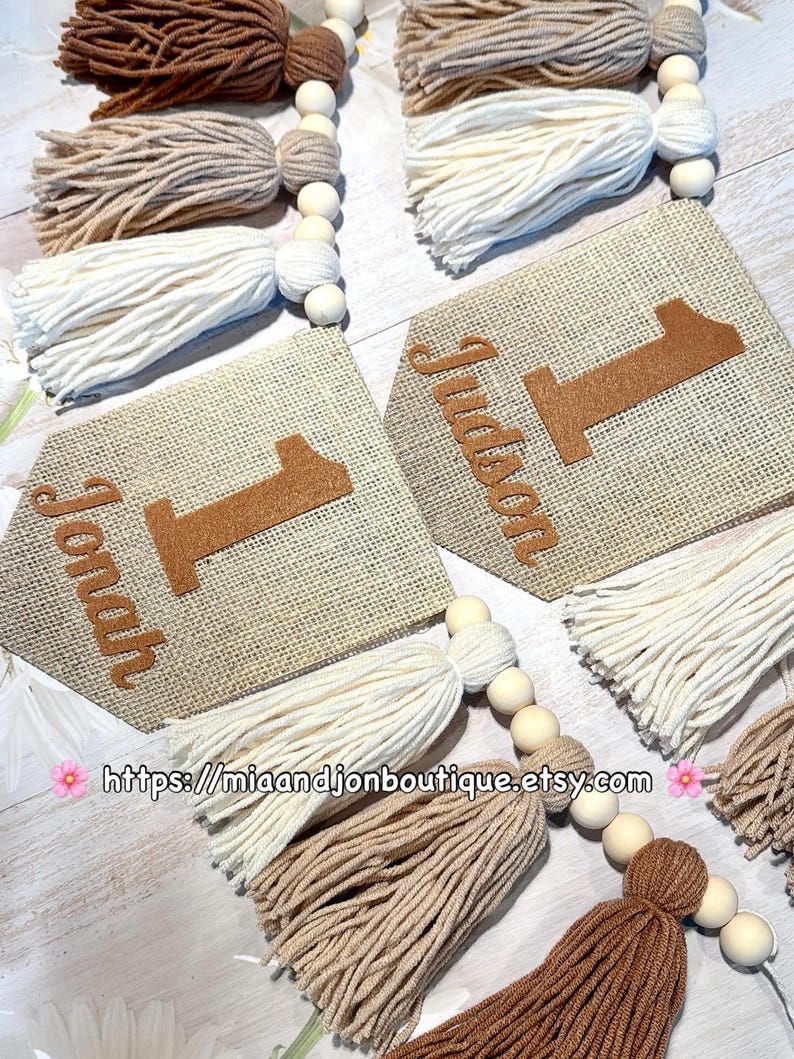 May include: Two burlap pennant banners featuring the names Jonah and Judson with the number one in brown felt. The banners are accented with strings of wooden beads and yarn tassels in shades of cream, beige, and brown. Text reads https://miaandjonboutique.etsy.com.