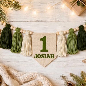May include: A decorative garland featuring a central burlap pennant with the number 1 and the name JOSIAH printed in dark green. The banner is strung with natural wooden beads and alternating dark green, olive green, and cream yarn tassels.