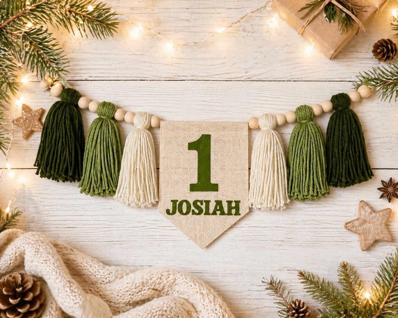May include: A decorative garland featuring a central burlap pennant with the number 1 and the name JOSIAH printed in dark green. The banner is strung with natural wooden beads and alternating dark green, olive green, and cream yarn tassels.