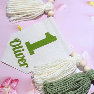 May include: A decorative garland featuring a cream-colored fabric pennant with the green text "Oliver" and the number "1" printed on it. The pennant is strung with natural wooden beads and alternating yarn tassels in olive green and cream.