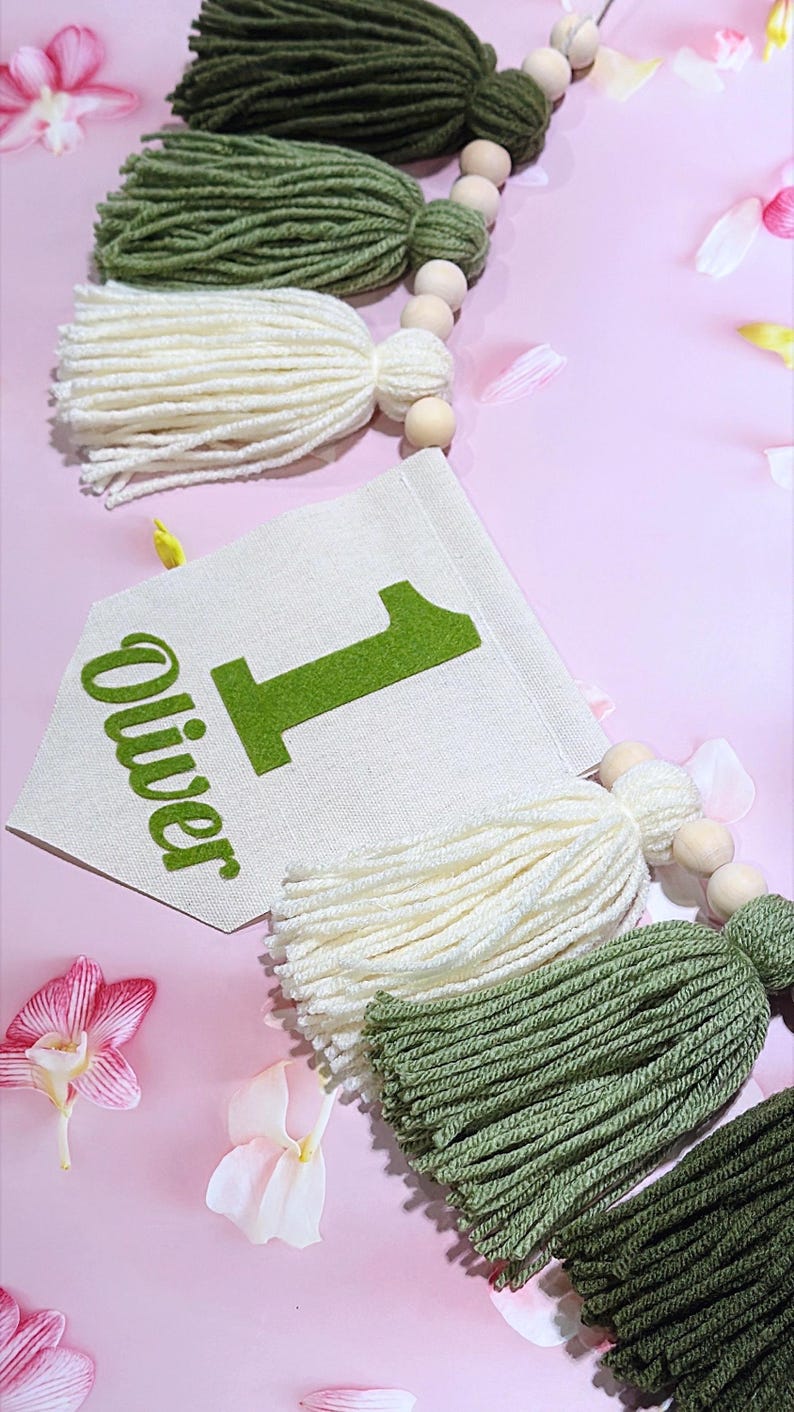 May include: A decorative garland featuring a cream-colored fabric pennant with the green text "Oliver" and the number "1" printed on it. The pennant is strung with natural wooden beads and alternating yarn tassels in olive green and cream.