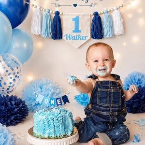 May include: A birthday decoration set featuring a banner with the text '1 Walker' and blue and white tassels, a blue cake topper with the text 'ONE', blue balloons, and blue paper pom-poms. The banner includes wooden beads and a heart icon.