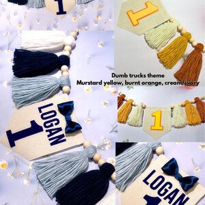 May include: A decorative banner featuring fabric pennants with the name LOGAN and the number 1, accented by a navy blue satin bow. The banner includes wooden beads and yarn tassels in shades of navy, light blue, white, mustard, and burnt orange.