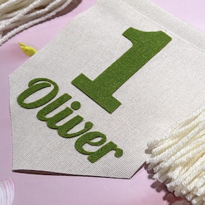 May include: A cream-colored fabric pennant banner featuring a large green felt number one and the name Oliver in green felt lettering. The banner is accented with a string of wooden beads and decorative yarn tassels in olive green and cream.