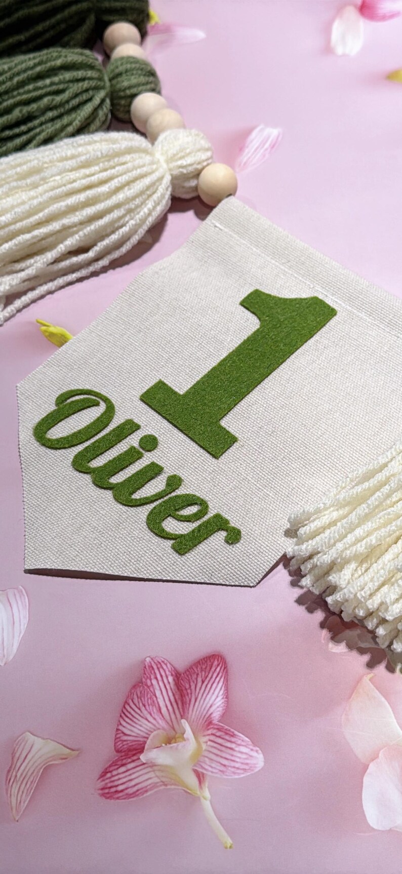 May include: A cream-colored fabric pennant banner featuring a large green felt number one and the name Oliver in green felt lettering. The banner is accented with a string of wooden beads and decorative yarn tassels in olive green and cream.