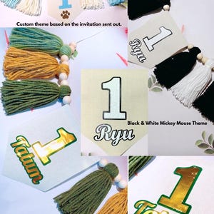 May include: A collection of decorative banners featuring the number 1 and various names like Ryu and Tatum. The banners are adorned with yarn tassels in colors including light blue, white, black, green, and gold, accented by wooden beads.