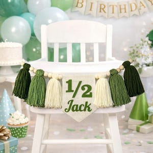 May include: A high chair banner featuring a central burlap pennant with the green text 1/2 Jack, flanked by alternating dark green, light green, and cream-colored yarn tassels strung on a wooden bead garland for a birthday celebration.