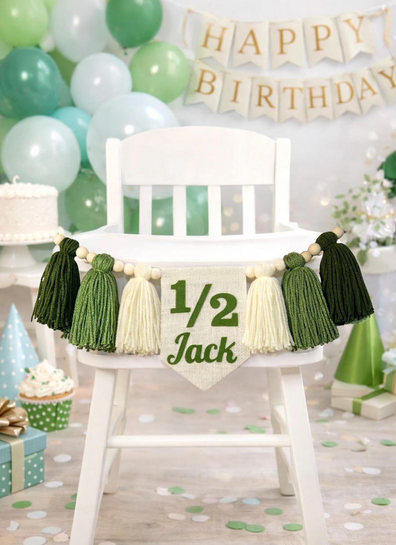 May include: A high chair banner featuring a central burlap pennant with the green text 1/2 Jack, flanked by alternating dark green, light green, and cream-colored yarn tassels strung on a wooden bead garland for a birthday celebration.