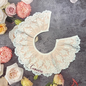 May include: A light pink baby bib with white floral lace trim and the name "Geneva" embroidered in red. The bib is surrounded by flowers in shades of pink, white, and yellow on a gray surface.