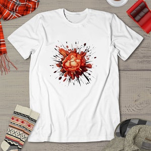 May include: White t-shirt featuring a graphic design of a red and orange explosion with black splatters. The explosion is centrally positioned on the shirt. The shirt is displayed on a wooden surface with a red and black plaid scarf, patterned socks, and a gray coat partially visible in the image.