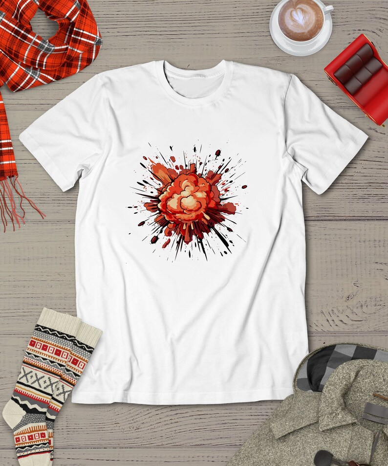 May include: White t-shirt featuring a graphic design of a red and orange explosion with black splatters. The explosion is centrally positioned on the shirt. The shirt is displayed on a wooden surface with a red and black plaid scarf, patterned socks, and a gray coat partially visible in the image.