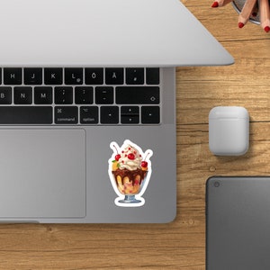 May include: A vibrant sticker featuring a delectable dessert: a banana split brimming with layers of creamy vanilla ice cream, rich chocolate sauce, and juicy red cherries. The banana split is elegantly presented in a clear glass dish, adding to its visual appeal. The sticker is a delightful representation of a classic sweet treat, perfect for adding a touch of whimsy to laptops, notebooks, or other smooth surfaces.