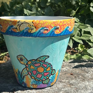 May include: A hand-painted terracotta plant pot featuring a light blue base with a colourful sea turtle illustration. The rim displays a vibrant sunset scene with orange, yellow, and blue waves. The bottom section includes painted coral reef details.