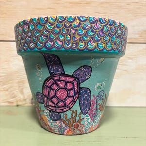 Könnte beinhalten: Handbemalter tealfarbener Blumentopf mit einem lila und rosa Meeresschildkröten-Design. Der Rand des Topfes hat ein Schuppenmuster in Gold, Teal und Lila. Korallen und Blasen sind ebenfalls auf den Topf gemalt.