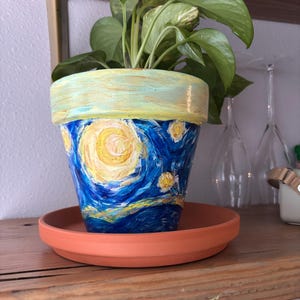 Può includere: Un vaso di fiori dipinto a mano con un design ispirato a "Notte stellata" di Van Gogh. Il vaso ha uno sfondo blu con stelle gialle e un grande sole giallo. È posto su un sottovaso in terracotta e contiene una pianta verde.