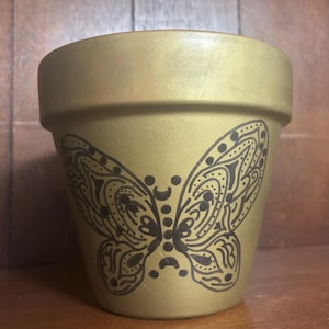 May include: A gold-colored ceramic flower pot with a black butterfly design. The butterfly is centered on the pot and has intricate patterns. The pot has a classic, rounded shape, suitable for planting flowers or herbs.