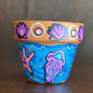 Puede incluir: Una pequeña maceta de terracota pintada a mano con un tema oceánico. La maceta presenta una base azul con un diseño de estrella de mar y medusa. El borde está decorado con patrones de conchas marinas y círculos en rosa, morado y blanco.