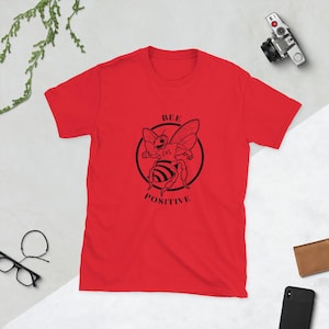 May include: A red t-shirt with a black bee illustration inside a circle. The words "BEE POSITIVE" are printed around the bee. The t-shirt is short-sleeved and made of a soft material.