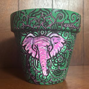May include: Hand-painted green ceramic flower pot featuring a pink elephant design. The pot is adorned with black and pink decorative patterns. The elephant is centered on the pot, with intricate details and a vibrant color palette.
