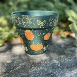 Könnte beinhalten: Handbemalter teal-farbener Keramik-Blumentopf mit orangefarbenen Kürbissen und goldenen Akzenten. Der Topf hat ein dekoratives goldenes Schleifen- und Wirbeldesign am Rand. Der Topf steht auf einer grauen Steinoberfläche.